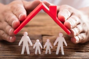 Die Hand einer Person schützt Familienfiguren mit rotem Dach auf einem Holzschreibtisch