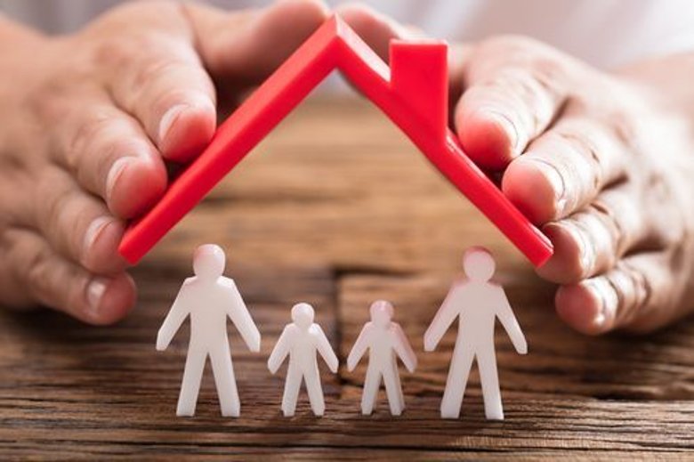Person schützt Familienfiguren mit Dach Die Hand einer Person schützt Familienfiguren mit rotem Dach auf einem Holzschreibtisch