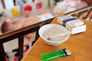 Aschenbecher mit angesteckter Zigarette auf einem Holztisch vor einem Babybett, grünes Feuerzeug, Zigarettenschachtel