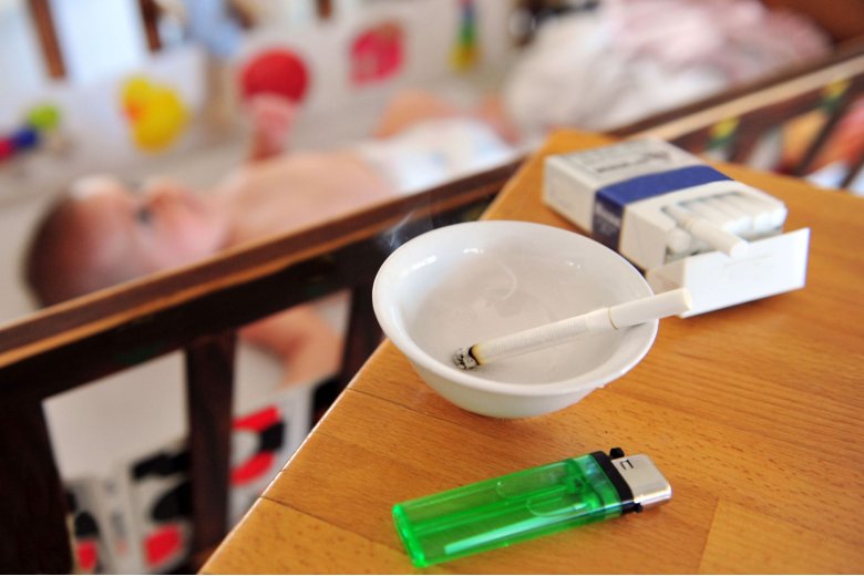 Rauchen in der Nähe von Kindern Aschenbecher mit angesteckter Zigarette auf einem Holztisch vor einem Babybett, grünes Feuerzeug, Zigarettenschachtel