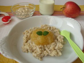 buntes Platzdeckchen, Babyrassel, Apfel, Schale mit Müsli und einem Glas Milch, sowie einen Teller mit Haferbrei und einem Klaks Apfelkompott und Minzblättchen und hellgrünen Plastiklöffel