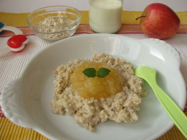 buntes Platzdeckchen, Babyrassel, Apfel, Schale mit Müsli und einem Glas Milch, sowie einen Teller mit Haferbrei und einem Klaks Apfelkompott und Minzblättchen und hellgrünen Plastiklöffel