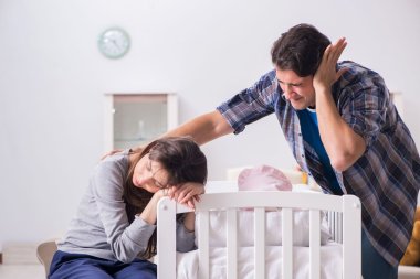 Vater hält sich das Ohr zu, Mutter ist erschöpft, schreiendes Baby liegt in einem weißen Bett