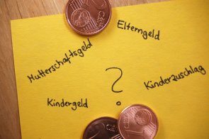 Gelber Zettel auf einer Holzplatte mit Centmünzen und den Begriffen Mutterschaftsgeld, Elterngeld, Kindergeld und Kinderzuschlag