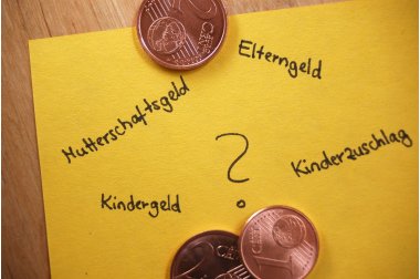 Elterngeld Gelber Zettel auf einer Holzplatte mit Centmünzen und den Begriffen Mutterschaftsgeld, Elterngeld, Kindergeld und Kinderzuschlag