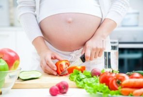 Schwangere Frau schneidet Tomaten, auf dem Tisch liegen weitere Zutaten für einen Salat