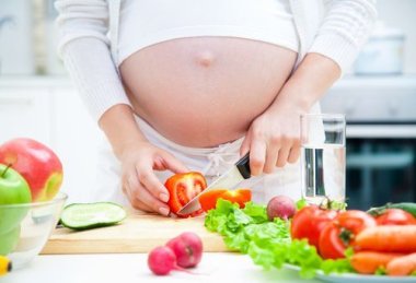 Schwangere Frau in der Küche macht Salat Schwangere Frau schneidet Tomaten, auf dem Tisch liegen weitere Zutaten für einen Salat