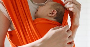 Gesichtslose Aufnahme einer Mutter, die ein schlafendes Neugeborenes in einem roten Tragetuch trägt und es mit einer zarten