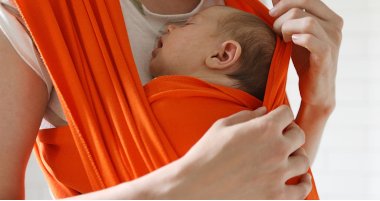 Frau trägt Neugeborenes in Babyschlaufe Gesichtslose Aufnahme einer Mutter, die ein schlafendes Neugeborenes in einem roten Tragetuch trägt und es mit einer zarten