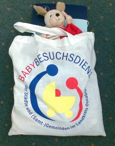 Tasche Babybesuchsdienst dunkler Hintergrund weisse Tasche mit Inhalt Stofftier Aufschrift Babybesuchsdienst stilisierter gelber Kinderwagen umrahmt von einer blauen und roten Figur die sich über den Kinderwagen beugen