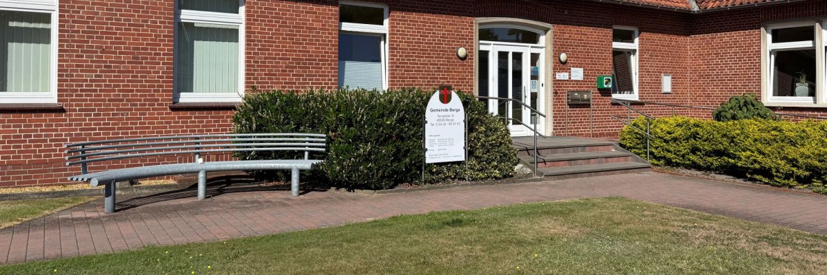 Gebäude mit weißen Fenster, Eingang über eine Treppe, davor eine eiserne Bank, ein Hinweisschild, Sträucher und Rasenfläche