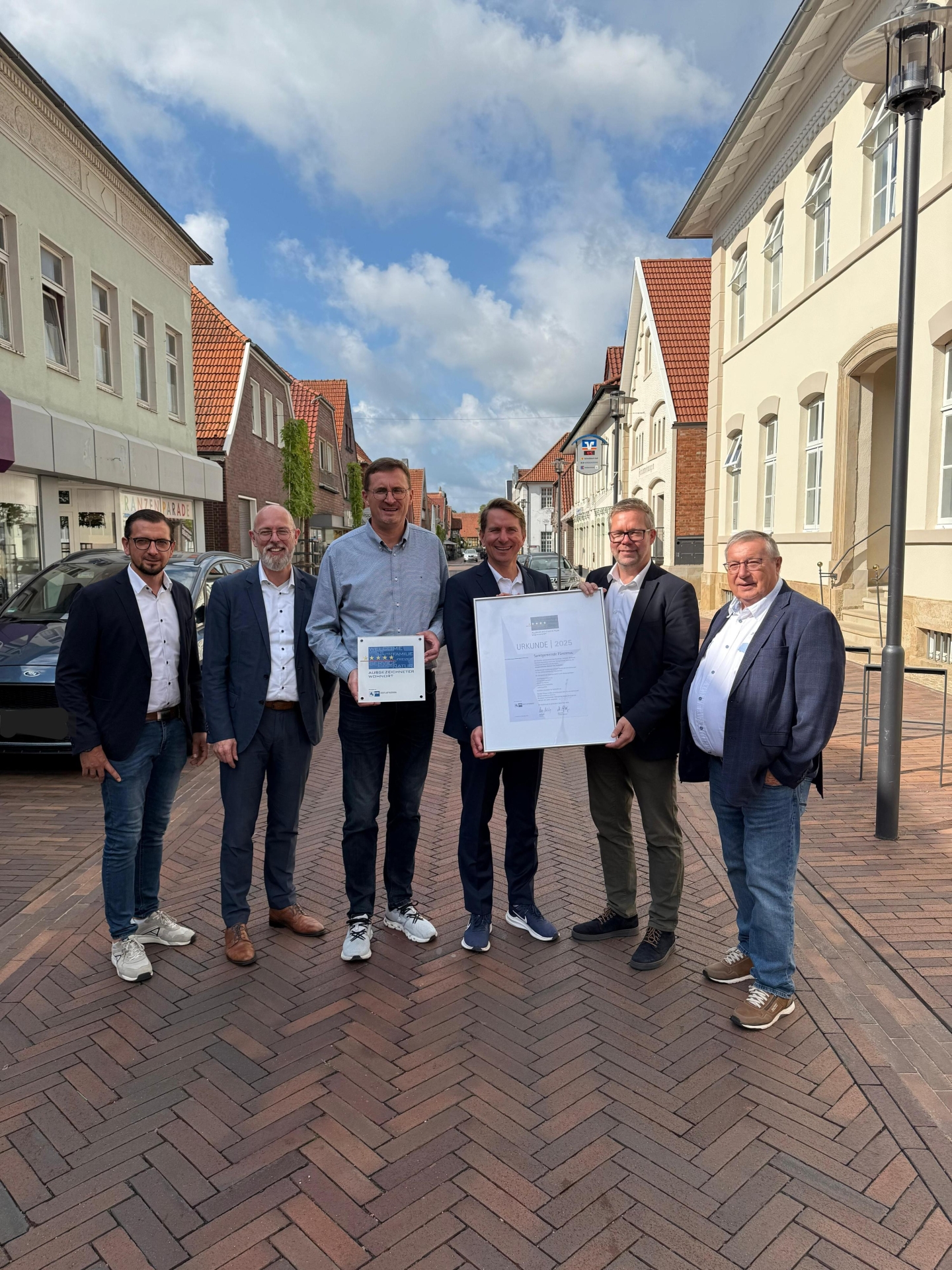 6 männliche Personen stehen auf einer rot geplasterten Straße in der Innenstadt, links und rechts sind Häuserzeilen, blauer wolkiger Himmel, es wird eine Urkunde überreicht