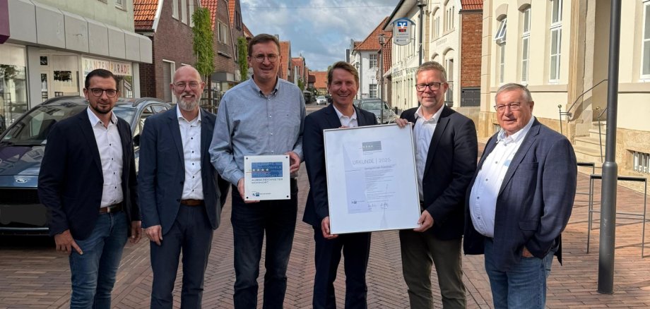 6 männliche Personen stehen auf einer rot geplasterten Straße in der Innenstadt, links und rechts sind Häuserzeilen, blauer wolkiger Himmel, es wird eine Urkunde überreicht