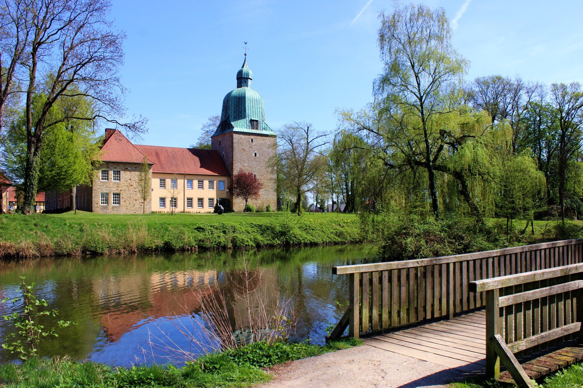 landschaftliches Frühlingsbild mit Schloßgebäude im Hintergrund, Schloßteich mit einer Brücke, Bäume