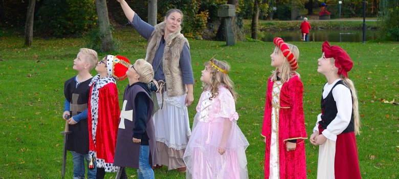Eine Frau zeigt 6 Kindern in mittelalterlicher Kleidung auf einer Wiese auf eine hohen Gegenstand, im Hintergrund ist eine Teich mit Bäumen, eine rotgekleidete Person